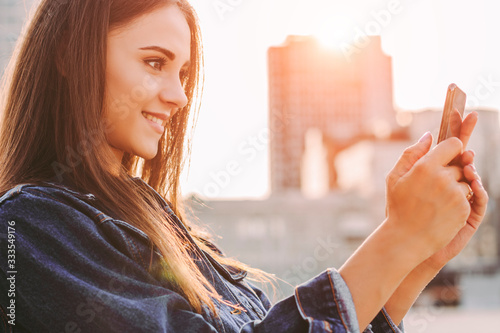 Happy attractive hipster girl taking photo on cell phone and smiling on city street. Beautiful trendy young woman using mobile phone to make selfie picture outdoors. Photographing for social networks