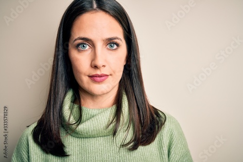 Young brunette woman with blue eyes wearing turtleneck sweater over white background with a confident expression on smart face thinking serious