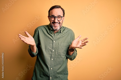 Middle age hoary man wearing casual green shirt and glasses over isolated yellow background clueless and confused expression with arms and hands raised. Doubt concept.