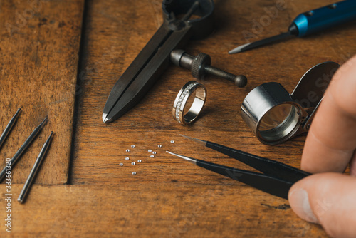 Jewelry craftsmanship. Jeweler holds tweezers. The jeweller engraver tools and golden ring, diamonds on wooden vintage desk. Tweezers, Magnifier, Chisel Hand Engraving, diamonds engagement ring