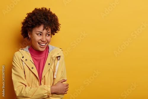 Half length shot of positive Afro American woman looks happily and carefree aside, wears casual jacket, poses against yellow background, blank space, hears hilarious story, has friendly conversation