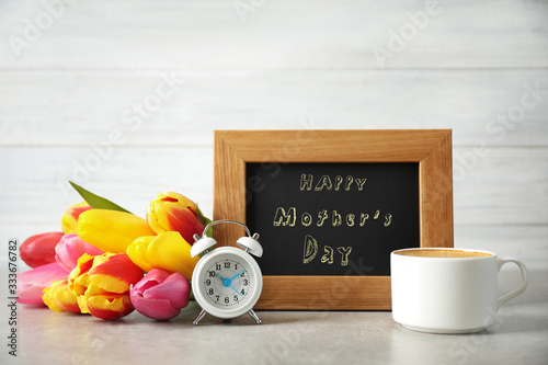 Chalkboard with phrase HAPPY MOTHER'S DAY, tulips, cup of coffee and alarm clock on light table