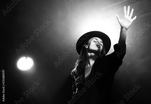 Beautiful braless actress girl, wearing an unbuttoned blazer and a hat, poses and dances in the warm light rays of scenic illuminators in the theatrical fog. Artistic, monochrome, noir design.