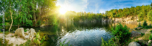 Panoramic landscape shot of idyllic lake surrounded by trees and cliffs, with the sun glowing on the horizon
