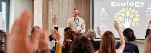 Developing leaders. Young male speaker answering questions while giving a talk at business meeting, ecological forum. Audience listening to him and raising hands to ask at the conference hall