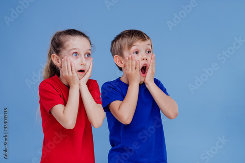 Astonished little girl and boy in casual clothes holding hands on faces and looking away while standing against blue background