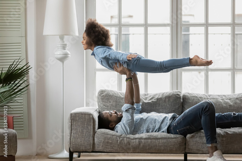 African dad lying on sofa playing with cute little daughter lift up her at stretched arms, happy kid girl imitates plane imagines herself like flying in air, activity at home and family travel concept