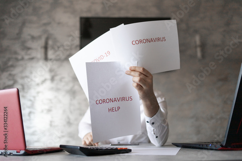 Pracownik w biurze trzymający kartki z napisami coronavirus, covid przed sobą. Biurko z laptopem, dokumetami i kalkulatorem. Coronavirus HELP