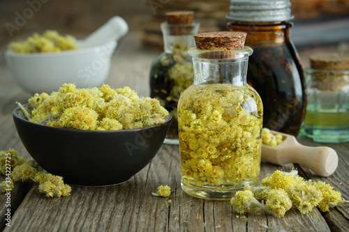 Helichrysum arenarium infusion bottle and dwarf everlast or immortelle dried flowers in bowl on wooden table. Tinctures and oil bottles on background.