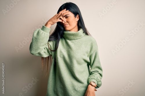 Young beautiful hispanic woman wearing green winter sweater over isolated background smelling something stinky and disgusting, intolerable smell, holding breath with fingers on nose. Bad smell