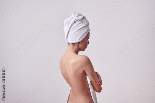 Skinny young woman after the shower from her back with white towel over her head, and with bare back. Half-length portrait against plain light grey background
