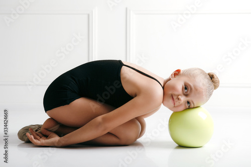 Young girl athlete rhythmic gymnastic in a black suit lyong on the floor, head on the ball, looking at camera smiling