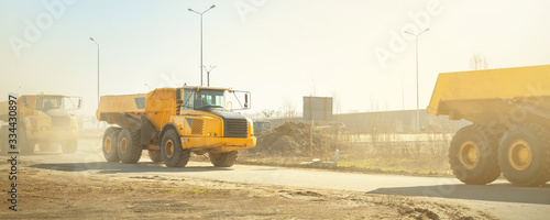 Many big articulated heavy industrial yellow dumper trucks driving on new highway road construction site on sunny day with blue sky background. Construction equipment machinery working on open pit