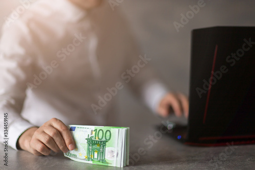 Nominały 100 Euro trzymane w dłoni przez mężczyzne przy laptopie. Koncepcja oferty bankowej, pożyczki, kredytu.