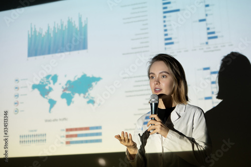 Young woman in lab coat gesturing hand and speaking into microphone while persuading investors in project importance