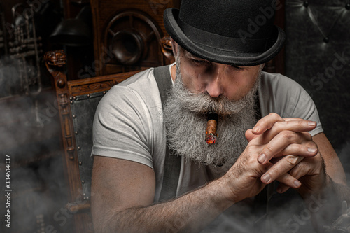 Old man smoking a cigar indoors. Mafia concept. Stylish male in his cabinet. Bearded man.