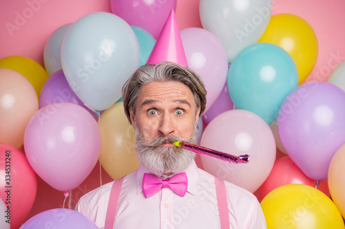 Close-up portrait of his he nice attractive cheerful funky grey-haired mature man having fun blowing festal whistle among colorful air balls isolated over pink pastel color background