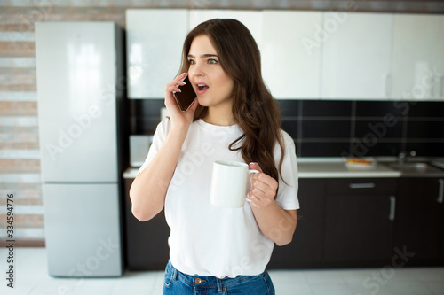 Young woman on kitchen during quarantine. Emotionally talking on phone and wonder. Hold white cup with drink in hand.