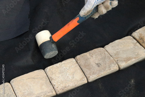 Worker's hand adjusting sandstone edging with a rubber mallet on the laid woven geotextile fabric in the summer garden under reconstruction