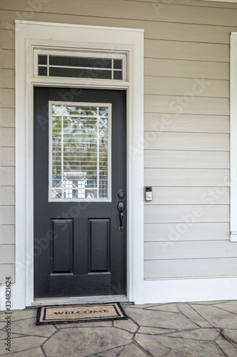 A black front door of a tan beige new construction house with siding. The door has a Transom window and a welcome mat.