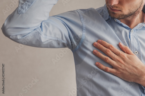 Man with hyperhidrosis sweating very badly under armpit in blue shirt, on grey.