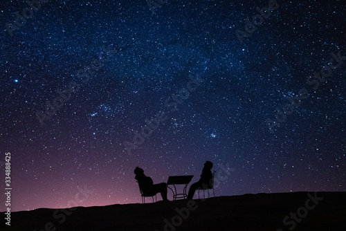 young couple sitting at a table on a desert dune while talking, relaxing and observing the stars and the milky way above them