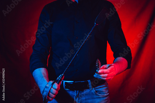 a man stands with a whip and an anal plug in his hands on a red background