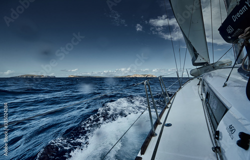 The view of the sea and mountains from the sailboat, edge of a board of the boat, slings and ropes, splashes from under the boat, sunny weather, dramatic sky