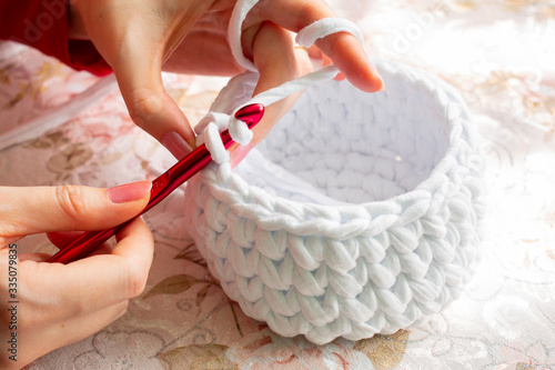 Young woman while crocheting on a patterned tablecloth, close up. Stay at home leisure activity idea. Basket made of white T-shirt yarn, with red crochet needle. Pale pink nails.