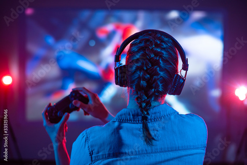 Girl in headphones plays a video game on a screen projector or TV in the dark room. Gamer with a joystick. Online games with friends, competitions. Fun entertainment. Teenagers play arcade. Back view