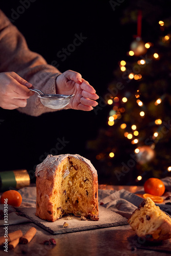 Hand Shaking Icing Sugar Over Christmas Panettone On Table Set For Meal With Tree Lights Behind