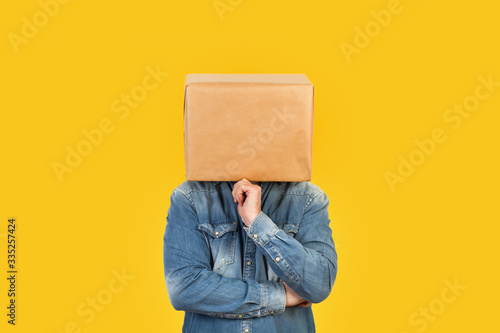 Hombre con caja de cartón en la cabeza con gesto pensativo y mano en la barbilla sobre fondo amarillo liso brillante aislado. Vista de frente. Copy space