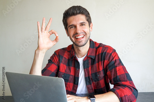 Young attractive male entrepreneur with a cute wide open smile sitting on the desc with his laptop showing OK sign with his fingers 