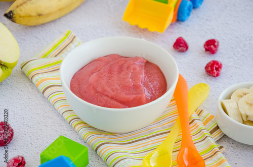 Baby food. Homemade apple puree or sauce with banana and raspberries in a bowl with a spoon on a light background. Healthy food. Horizontal orientation.