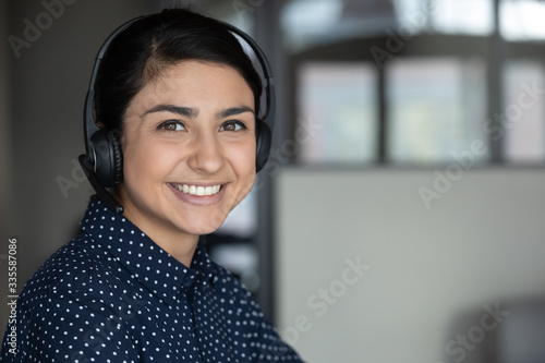 Head shot close up smiling attractive indian ethnicity millennial businesswoman wearing headphones, looking at camera. Happy sincere pleasant young mixed race female professional working remotely.