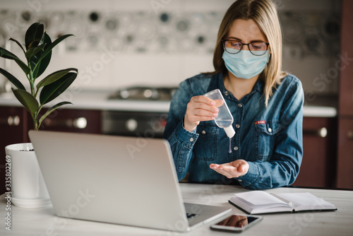 Coronavirus. Sanitizer gel. Business woman working, wearing protective mask in quarantine. Cleaning hands antibacterial gel to eliminate germs. Stay at home. Girl learns, using laptop. Freelance.