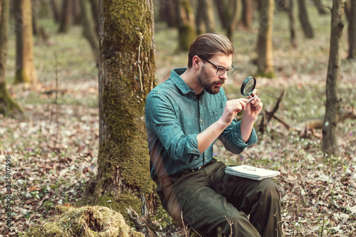 Concentrated botanist studying moss through magnifier holding it with tweezers sitting in forest