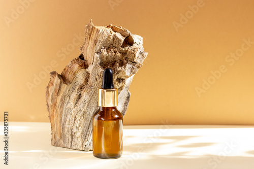 A glass cosmetic bottle with a dropper stands next to a bar of wood on a beige background with bright sunlight. The concept of natural cosmetics, natural essential oil