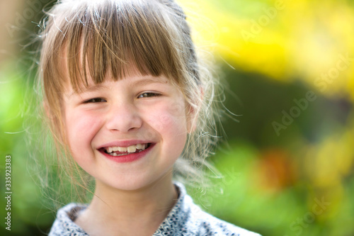 Cute pretty child girl with gray eyes and fair hair smiling in camera outdoors on blurred sunny green and yellow bright bokeh background.