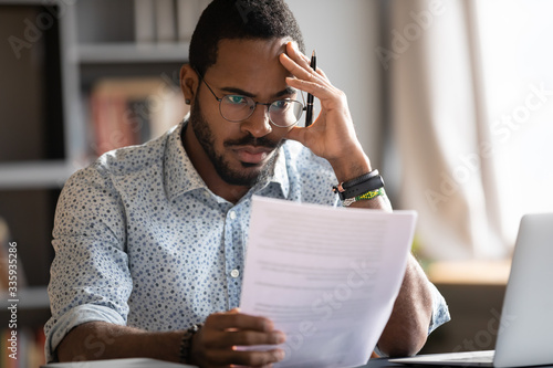 Pensive unhappy biracial man in glasses feel distressed reading bad news in paperwork letter, disappointed frustrated African American male confused by unpleasant postal correspondence or notice