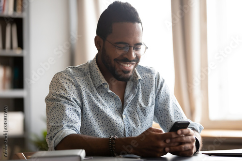 Happy biracial man in glasses sit at desk have fun texting messaging using wireless internet on smartphone gadget, smiling African American male in spectacles laugh watch funny video on cellphone