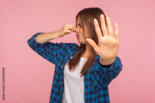 Unpleasant smell. Portrait of confused girl in checkered shirt grimacing in disgust and pinching her nose, displeased with bad breath, stinky odor, fart. indoor studio shot isolated on pink background