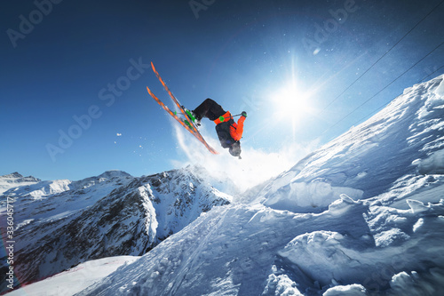 Low angle view athlete skier in an orange jacket does a back flip with flying powder of snow against a clear blue sky sun and snow-capped mountains of the Caucasus.