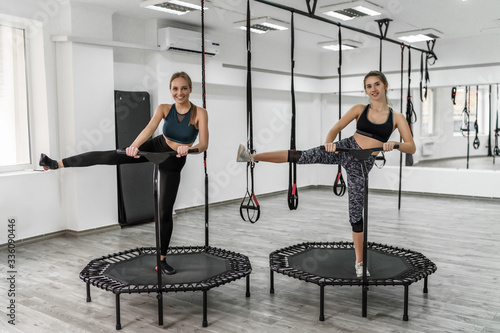 Portrait of two young pretty active girls in fitness clothes doing exercises for jump with trampoline during training in gym