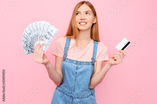 smiling young girl showing a lot of money in banknotes and holding a credit card isolated on a pink background