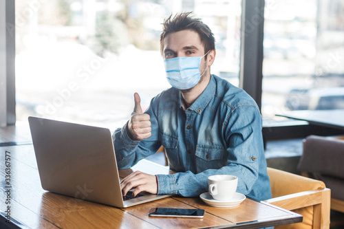 Everything alright! Portrait of satisfied young man with surgical medical mask in blue shirt are sitting and working on laptop, thumb up, looking at camera. indoor working and health care concept.