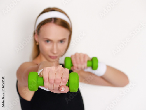 Beautiful young sporty woman in black outfit doing exercises with green dumbbells to stay healthy and active on self isolation in home quarantine posing against white wall focus on dumbbell