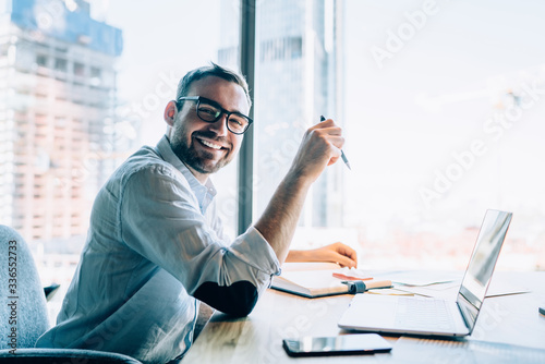 Portrait of cheerful male entrepreneur in classic eyewear smiling at camera while working at desktop table with modern laptop computer, happy executive manager in spectacles enjoying online marketing