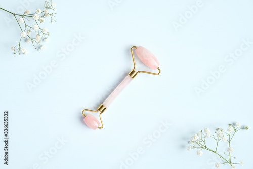 Rose quartz facial massage roller on blue background with gypsophila flowers. Facial massager with jade stone, anti-aging, anti-wrinkle beauty skincare tool