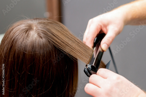 Hairdresser straightens hair of woman with hair straightener tool in hair salon.